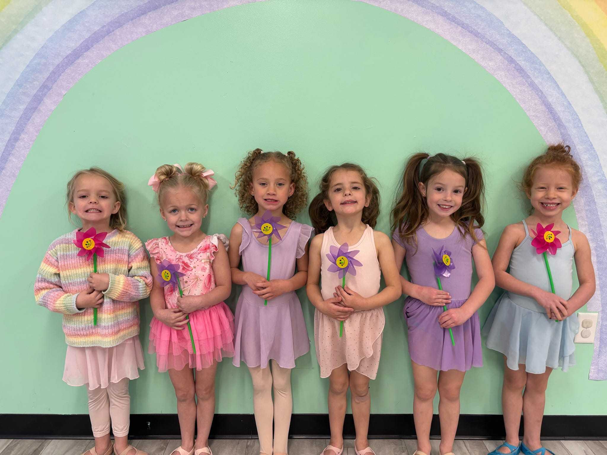 Six young E&E Dance Company dancers holding colorful flower pinwheels in front of the studio rainbow mural.