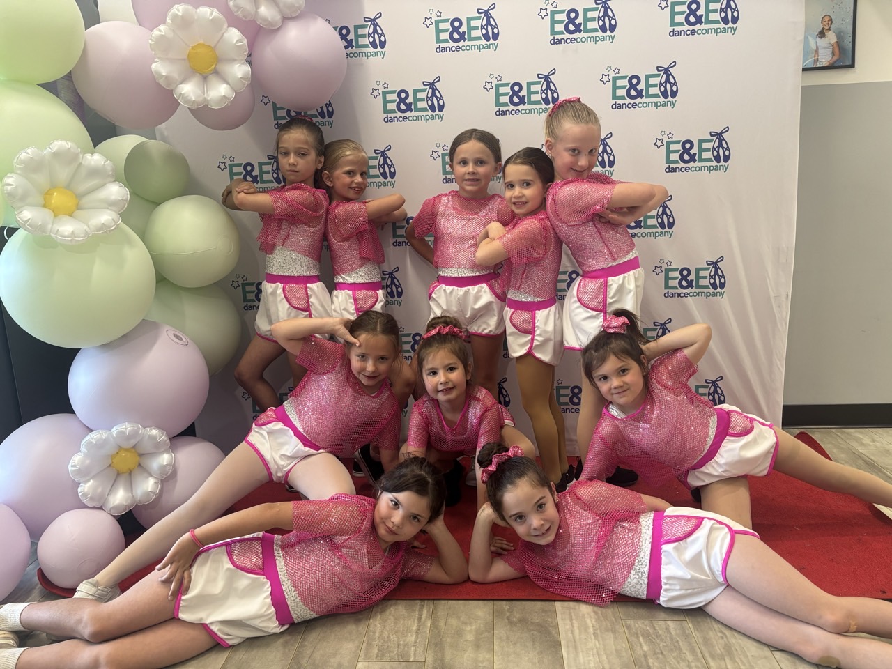 Young dancers in matching pink tops and white shorts posing in front of the E&E Dance Company recital backdrop.
