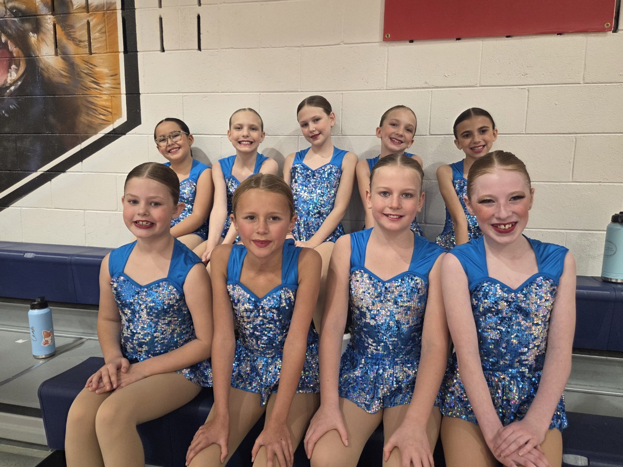 Collective dancers in blue sequined costumes smiling together on bleachers at the Fall Showcase.
