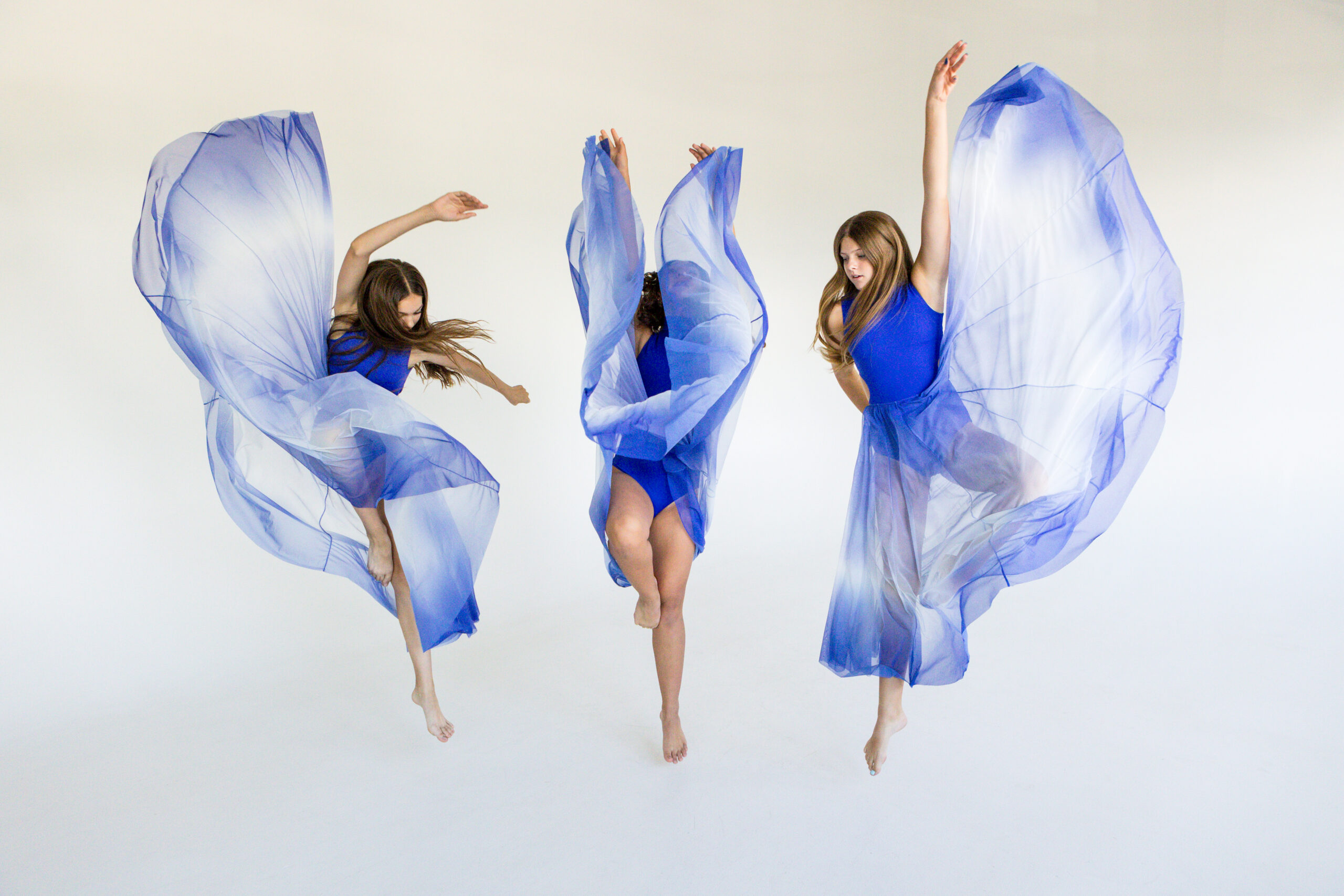 Three E&E Dance Company dancers in blue costumes lifting flowing skirts as they leap.